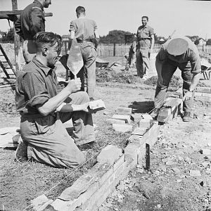 Guardsmen laying the first courses of brick to form the foundations of a nissen hut style temporary house; IWM D 25704